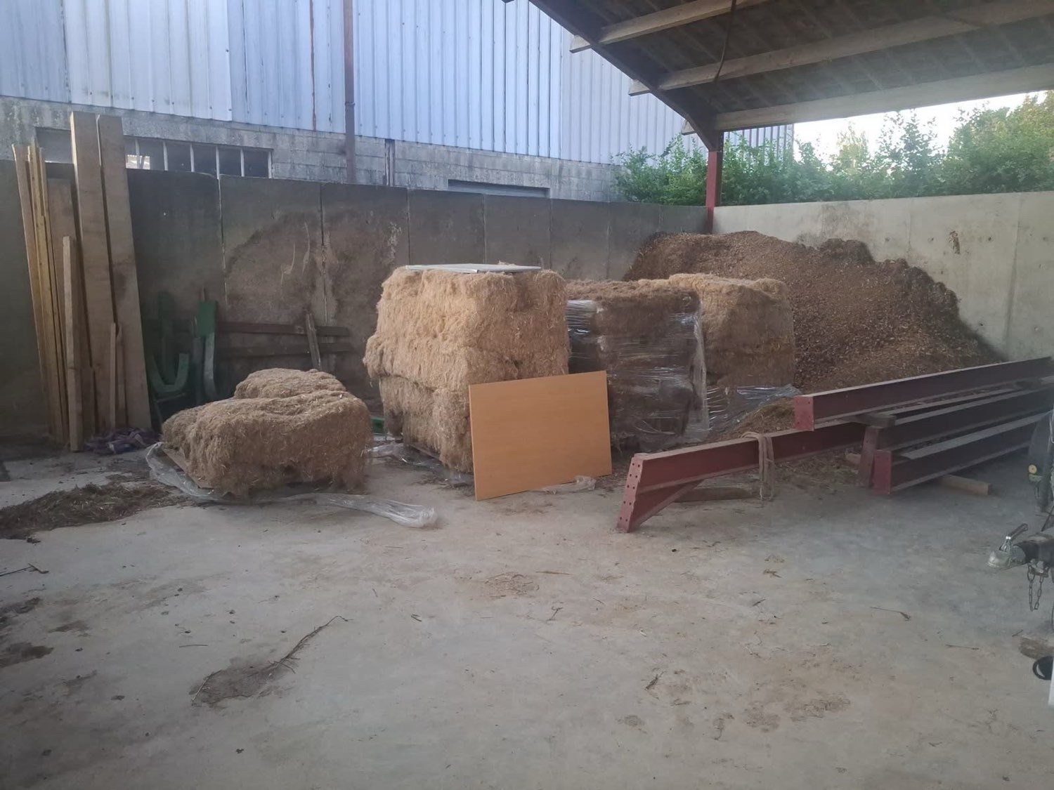 Bûches, plaquettes forestières et bottes de fourrage entassées sous un hangar agricole, avant passage en cellule de séchage